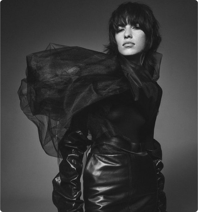 a black and white image of a woman wearing a leather skirt and black top with a sculptural tulle element on her neck and shoulder