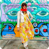 Woman in a yellow floral dress, white jacket, and red scarf standing in front of a colorful wall