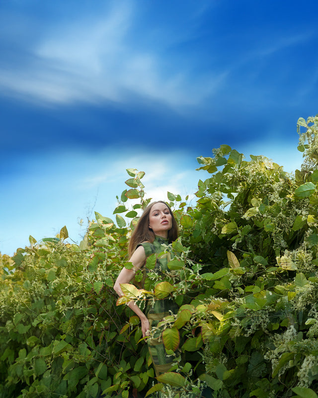 a thin white woman in a green dress standing in green plants with a bright blue sky above