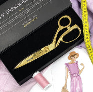 a photo of a pair of gold Mood branded fabric shears in a box with a spool of thread, tape measure, fabric swatches, and fashion sketches around it