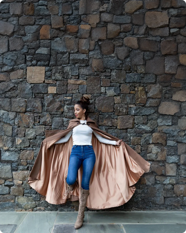 a woman in a light brown cape, white shirt, blue jeans and tan boots standing against a stone wall